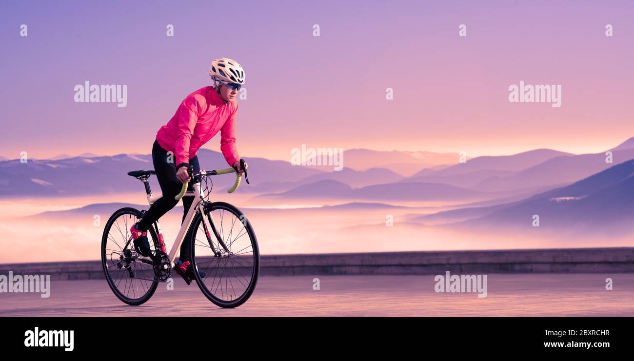 Junge Frau Radfahrerin Fahren Sie mit dem Fahrrad auf der Straße in den wunderschönen Bergen bei Purple Sunset. Abenteuer, Reisen, gesunder Lebensstil und Sportkonzept. Stockfoto
