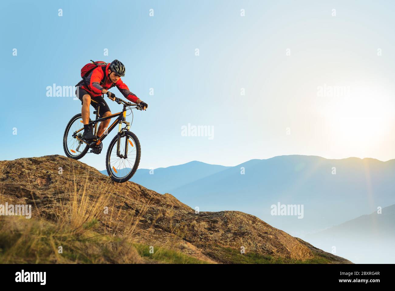 Radfahrer in roter Jacke mit dem Fahrrad in den schönen Bergen den Felsen hinunter auf dem Sunrise Sky Hintergrund. Extreme Sport und Enduro Biking Konzept. Stockfoto