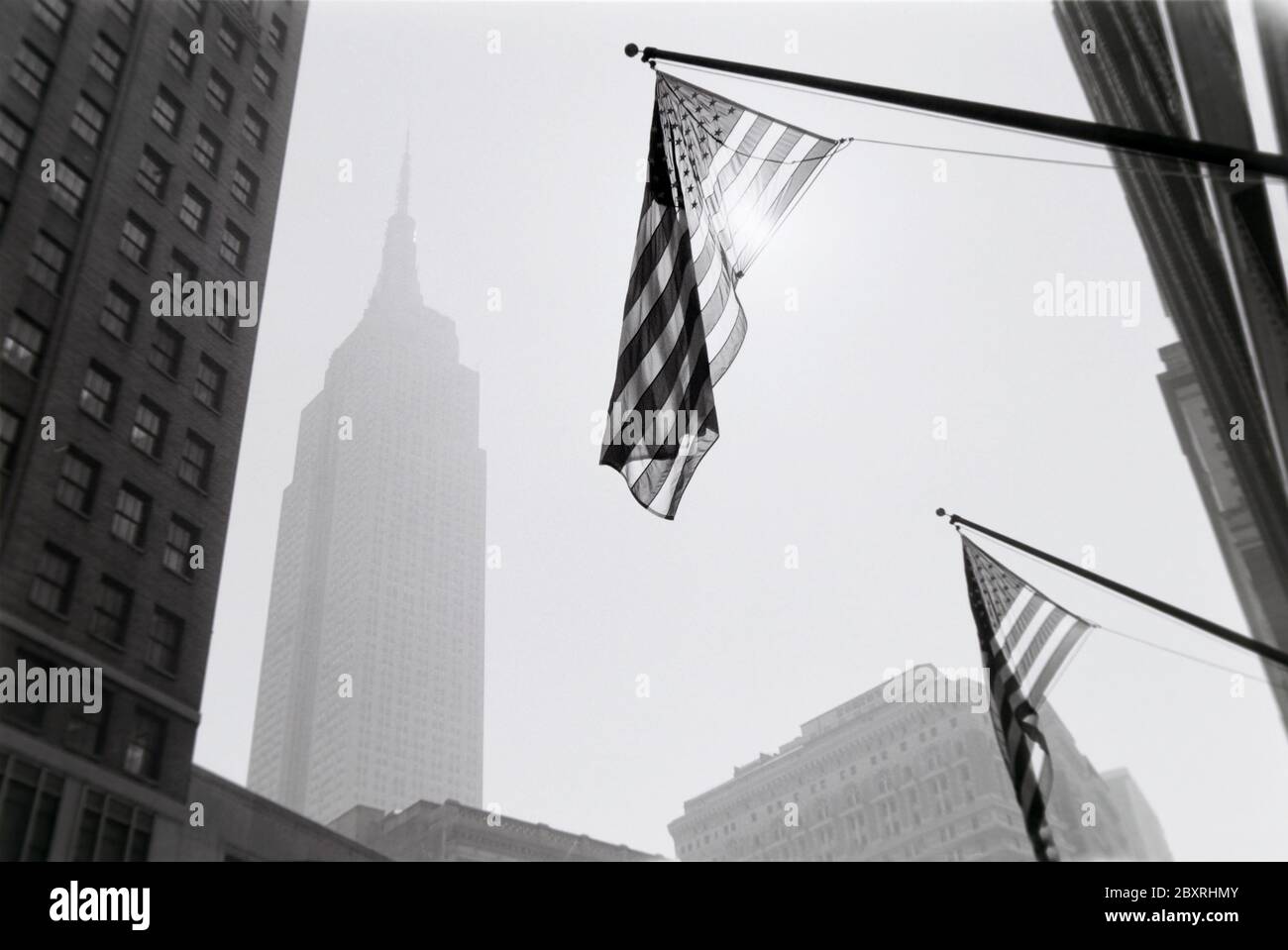 Schwarz-Weiß-Kunstfoto aus New York City mit US-Flaggen an Gebäuden, die von Sonnenreflexen und Empire State Building grau in der d Stockfoto