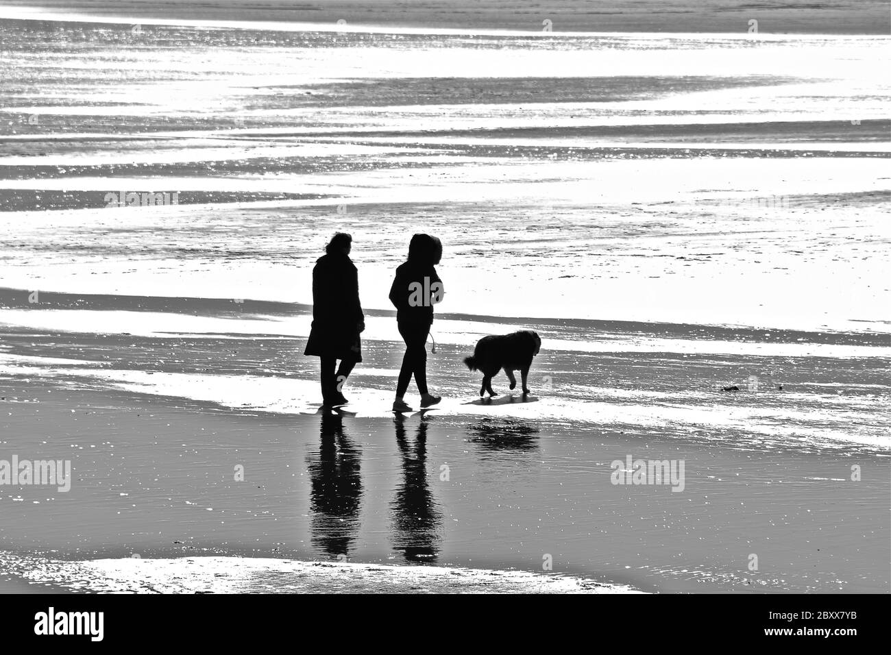 Ein schwarz-weißes Bild von zwei Menschen und ihrem Hund, die am Ufer am Strand von Sandy Bay, Porthcawl, spazieren gehen. Stockfoto