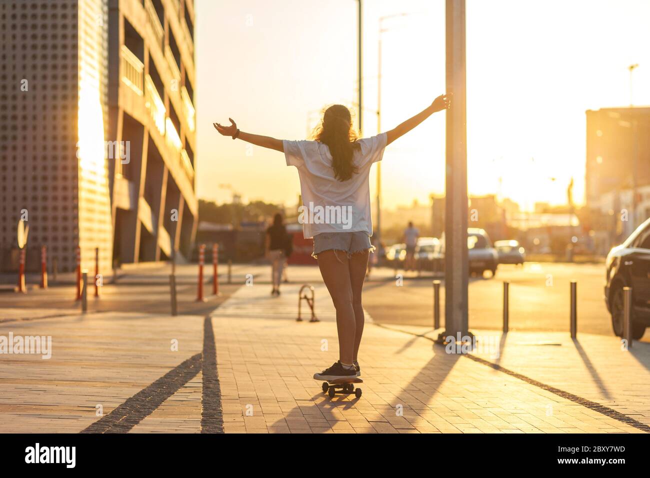 Mädchen auf einem Skateboard beleuchtet von den Strahlen der untergehenden Sonne. Fahrten auf einem Skateboard mit den Armen hoch, seinen Rücken zur Kamera angehoben. Selektiver Fokus. Sonnenuntergang in der Stadt. Hochwertige Fotos Stockfoto