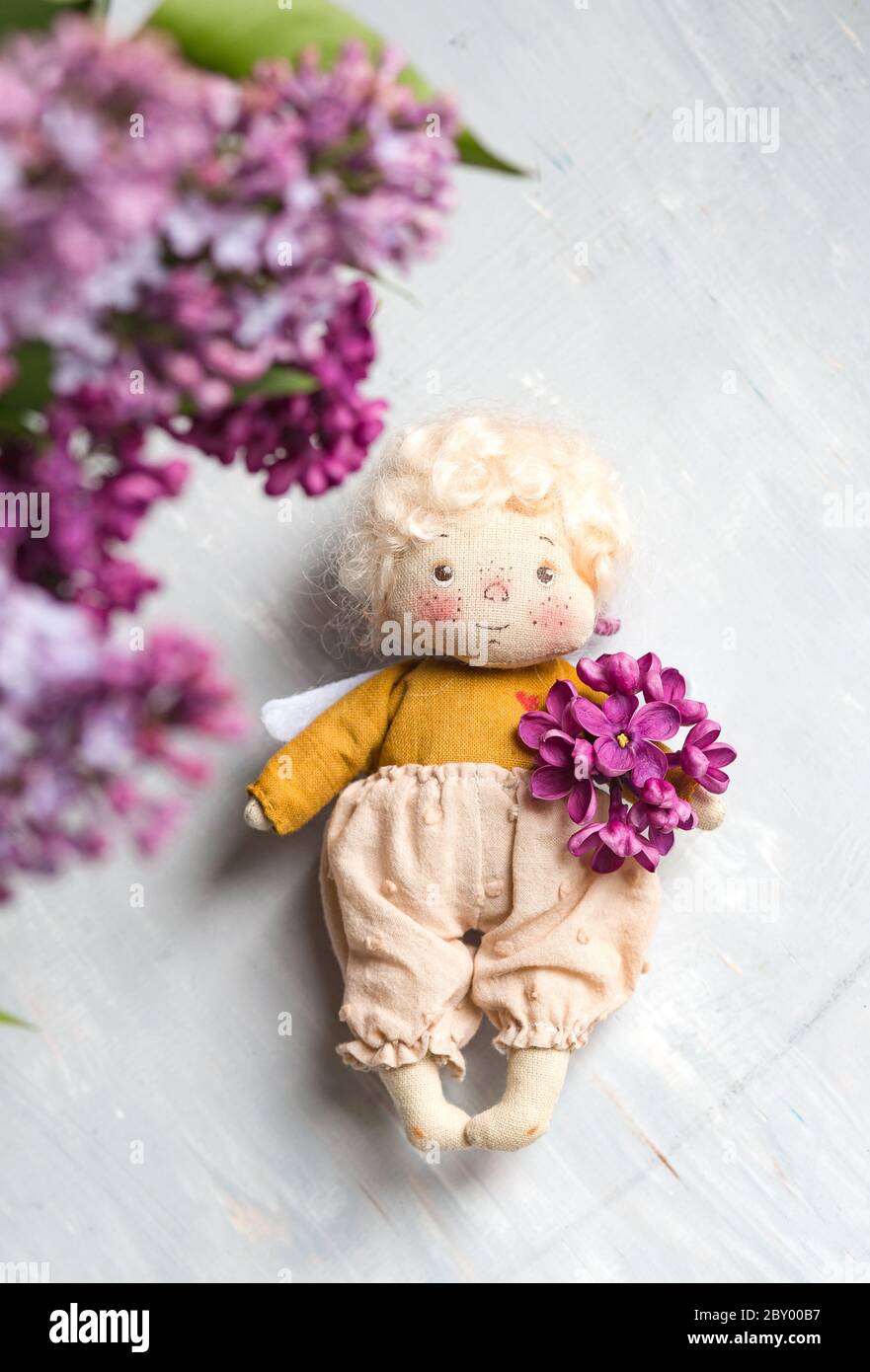 Kleiner goldhaariger Engel in den blauen, rosa, lila, violetten lila Blüten. Handgemachtes Spielzeug in violetten Flieder Farben. Stockfoto