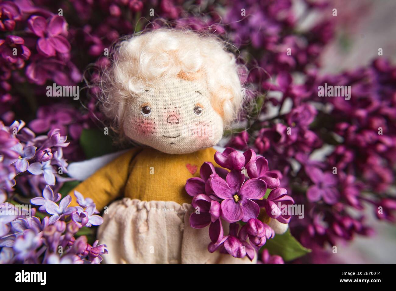 Kleiner goldhaariger Engel in den blauen, rosa, lila, violetten lila Blüten. Handgemachtes Spielzeug in violetten Flieder Farben. Stockfoto