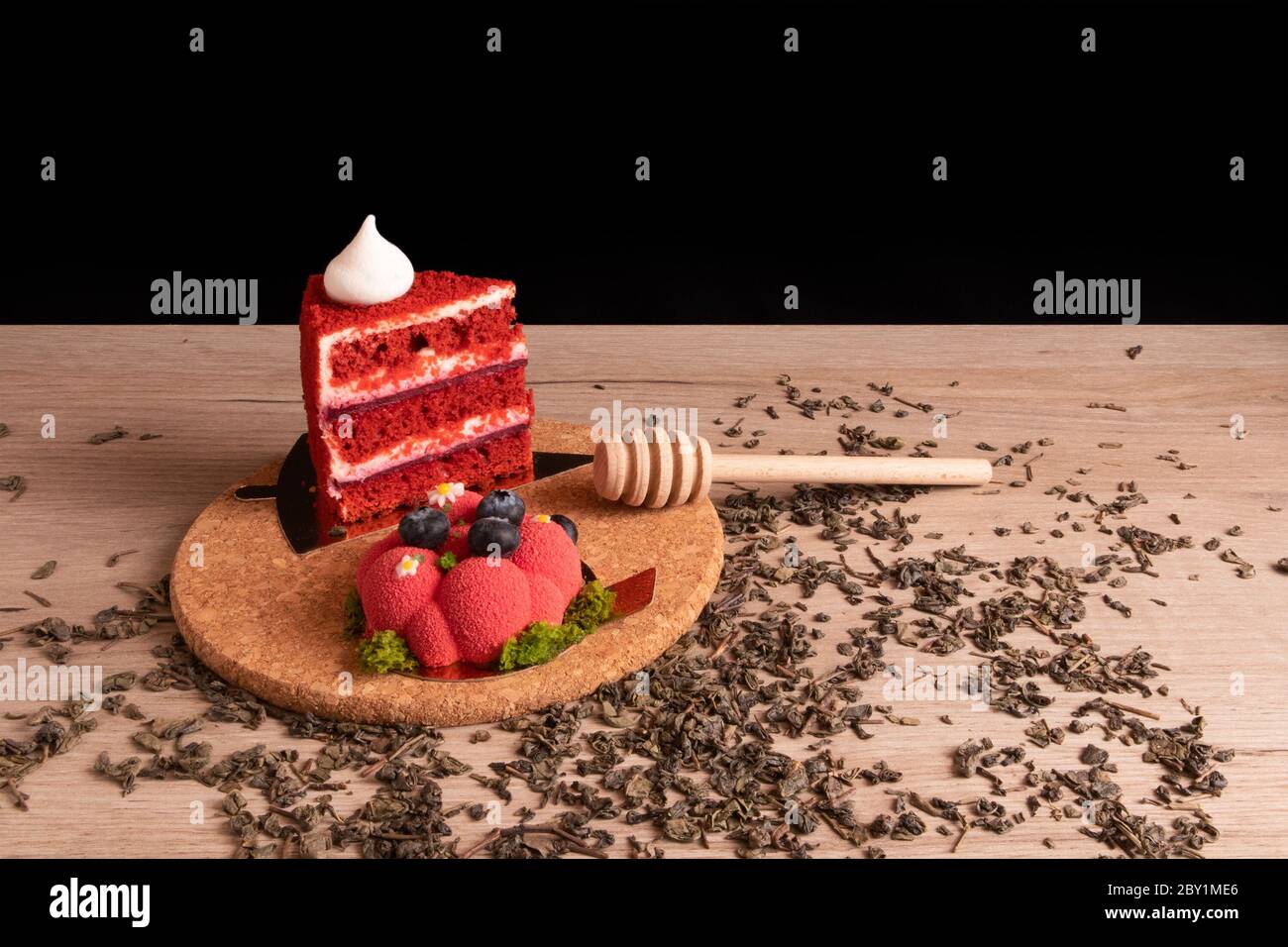 Zwei appetitlich leckere rote Kuchen und Honig Dipper auf einem Korkständer mit verstreuten trockenen grünen Tee auf einem Holzbrett Stockfoto