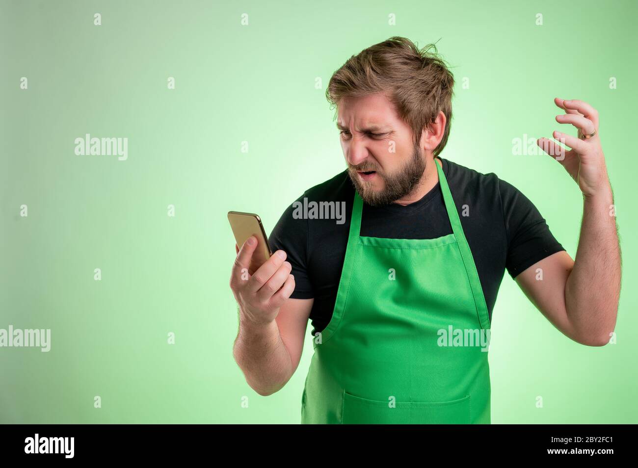 Supermarktmitarbeiter mit grüner Schürze und schwarzem T-Shirt, Smartphone haltend, SMS, nervös isoliert auf grünem Hintergrund Stockfoto