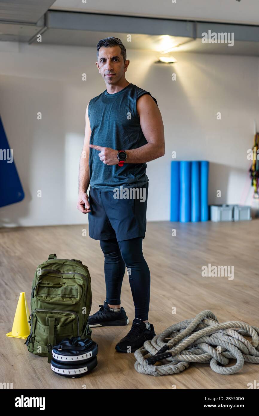 Motivierter Bootcamps-Trainer steht mit Fitnessgeräten in der Turnhalle. Hanteln, Wasserkocher, Seil, Sandsack auf Holzboden. Porträt des libanesischen inst Stockfoto