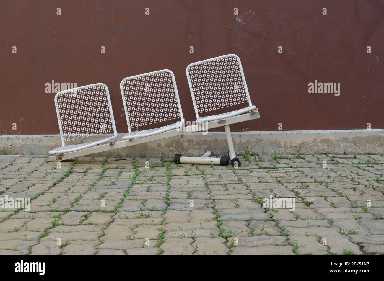 Reihe von beschädigten weißen Metallstühlen mit gebrochenem Bein, schrägen Stühlen auf Pflasterstein im Park verlassen Stockfoto