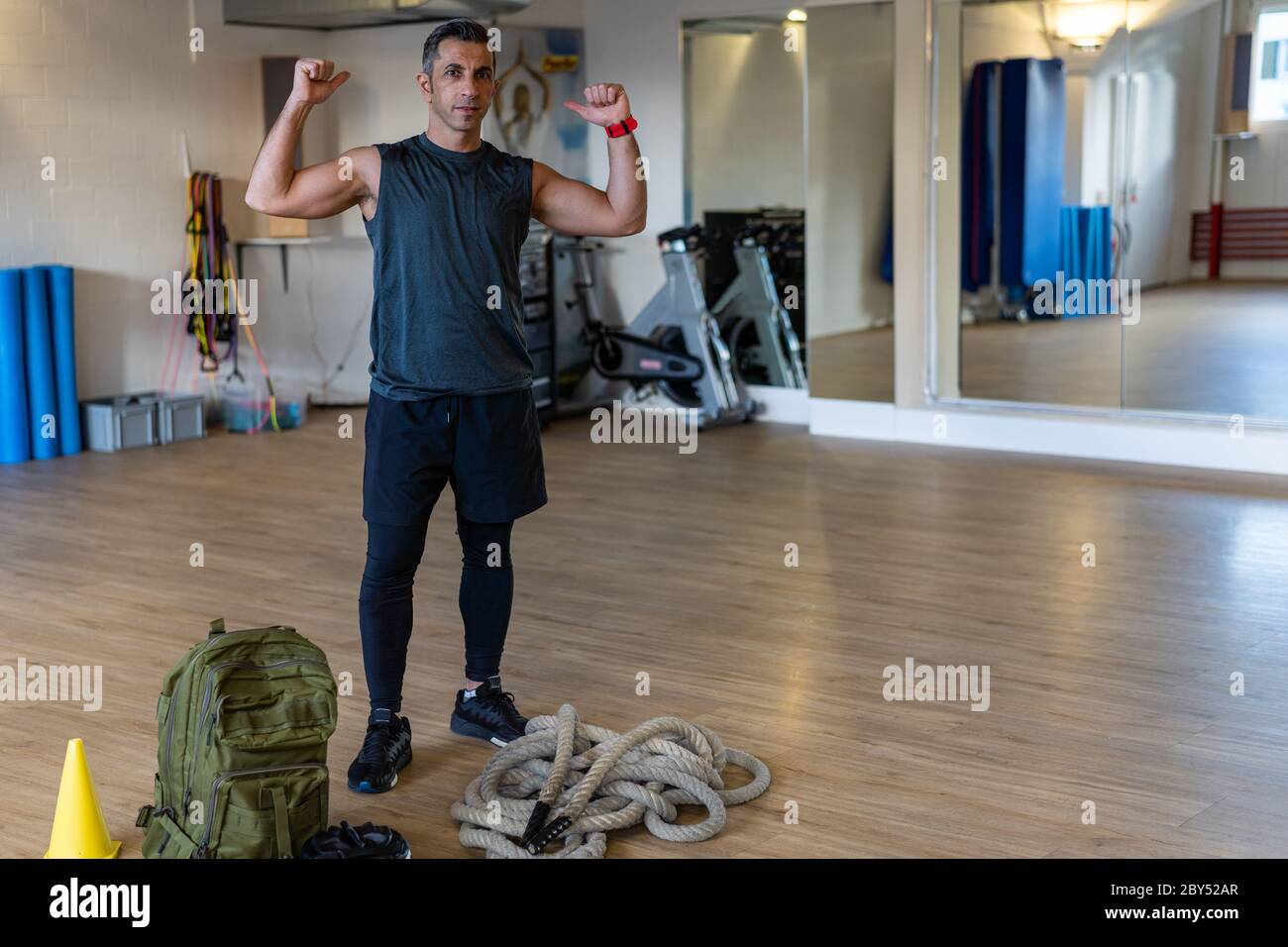 Motivierter Bootcamps-Trainer steht mit Fitnessgeräten in der Turnhalle. Hanteln, Seil, Sandsack auf Holzboden. Instruktor zeigt Muskeln. Hochformat f Stockfoto