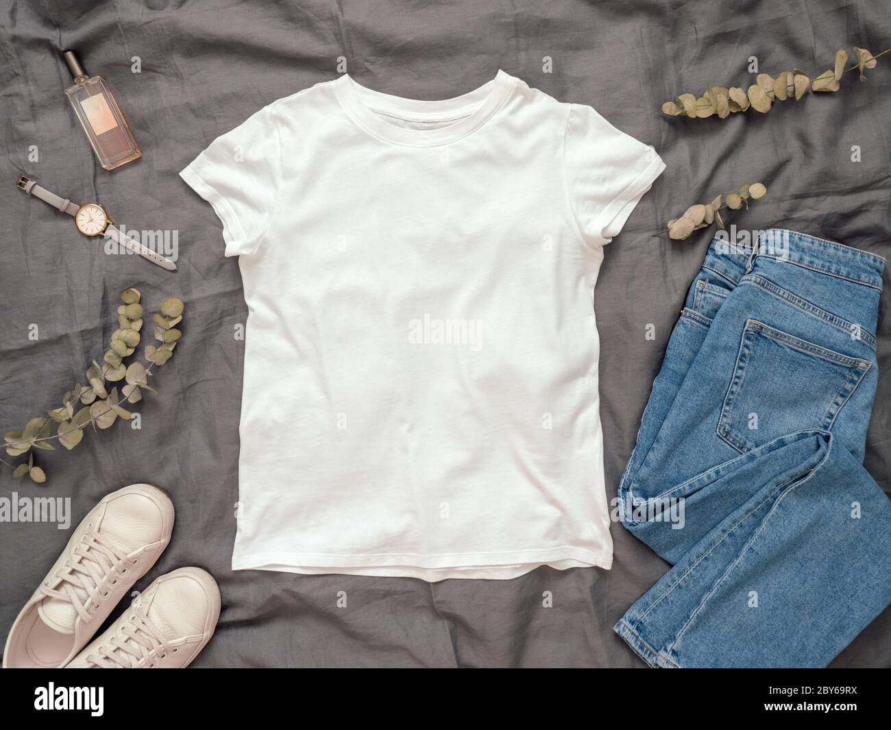 Modische Optik mit weißem leerem T-Shirt, Jeans und weißen Sneakers. Draufsicht des weißen, leeren T-Shirts mit kurzen Ärmeln über grauer Bettwäsche. Stehkragen für T-Shirt-Druck. Stockfoto