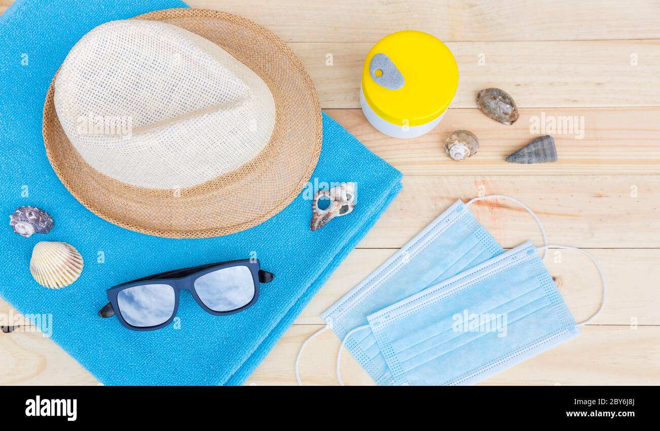 Draufsicht auf einige Strandartikel wie Handtuch, Hut und Sonnenbrille zusammen mit einem Paar Schutzmaske auf einem Holzbrett. Sommerferienkonzept Stockfoto