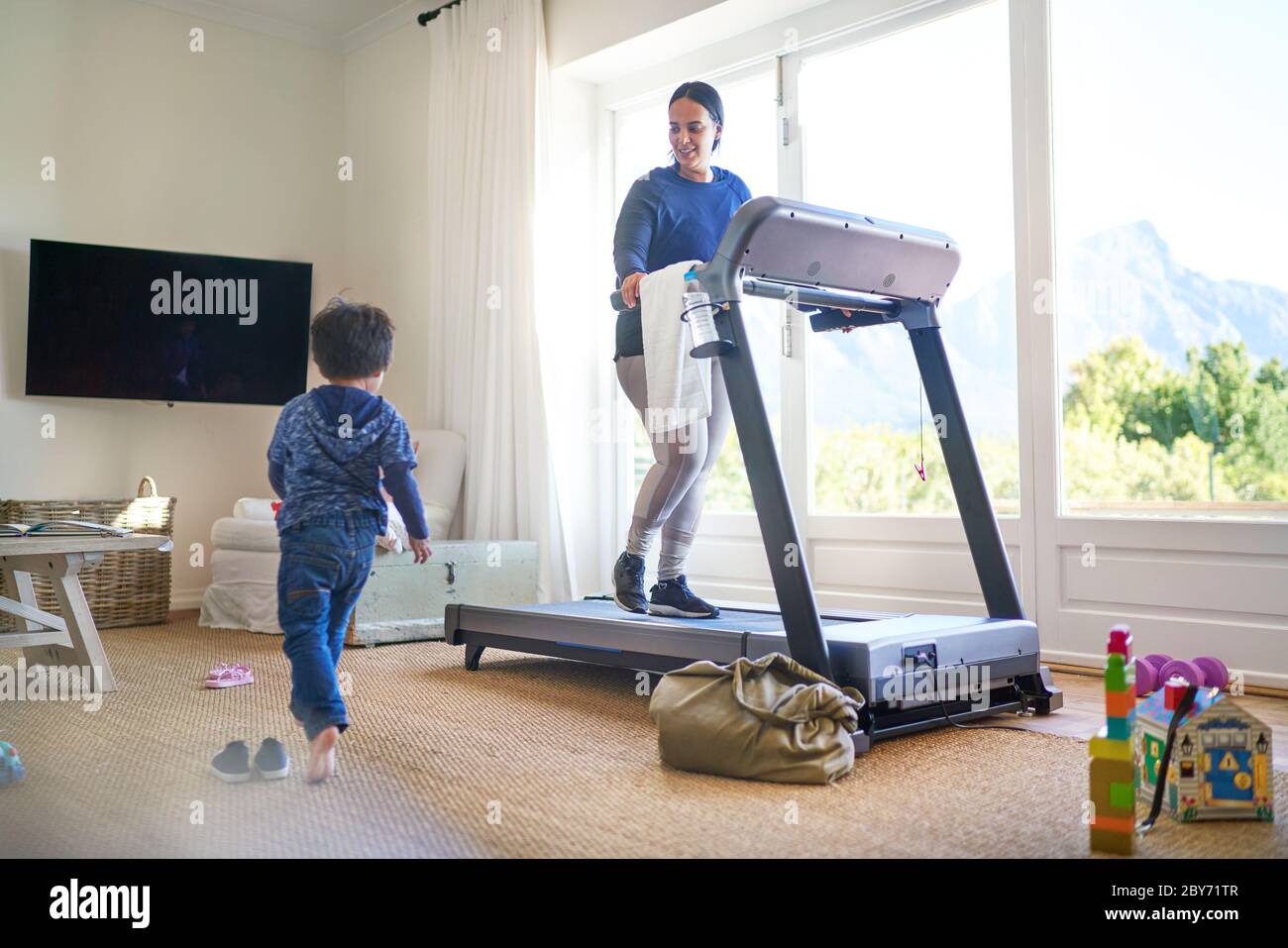 Junge spielt im Wohnzimmer, während Mama auf dem Laufband trainiert Stockfoto