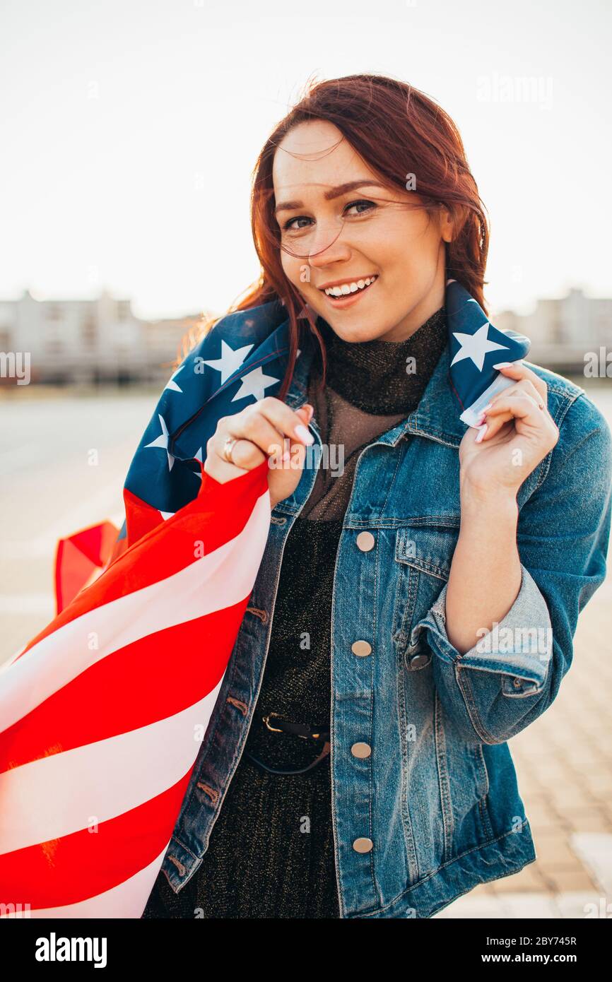 Junge hübsche rothaarige Frau mit nationaler us-Flagge über Sonnenschein gewickelt. Weiche Hintergrundbeleuchtung. Unabhängigkeitstag, amerikanischer Traum, Freiheitskonzept Stockfoto