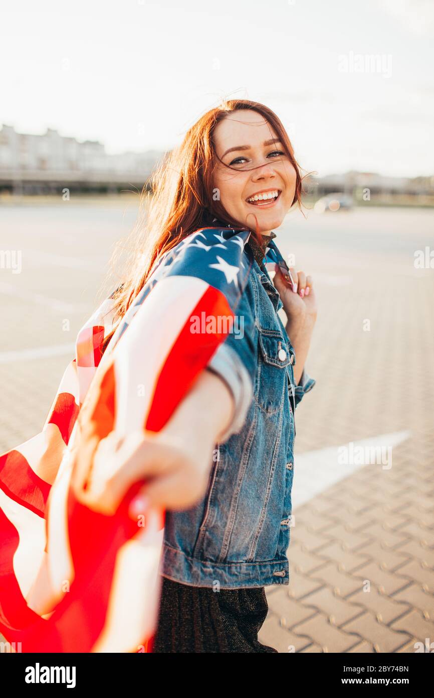 Junge hübsche rothaarige Frau mit nationaler us-Flagge über Sonnenschein gewickelt. Weiche Hintergrundbeleuchtung. Unabhängigkeitstag, amerikanischer Traum, Freiheitskonzept Stockfoto