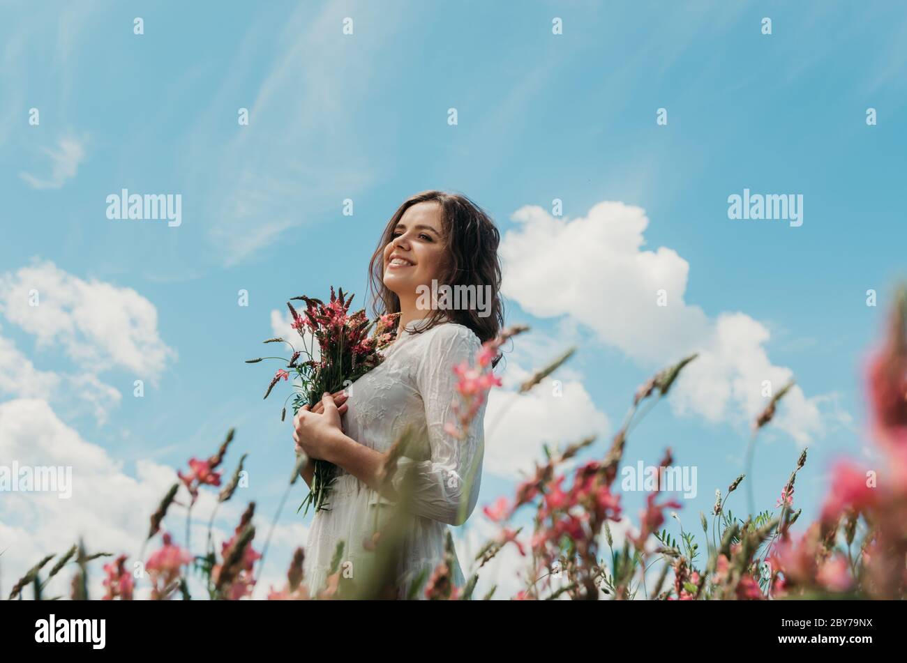 Mittelkörper Porträt einer jungen Frau in weißem Kleid lächelnd zur Seite und hält einen Blumenstrauß, gegen blauen hellen Himmel geschossen Stockfoto