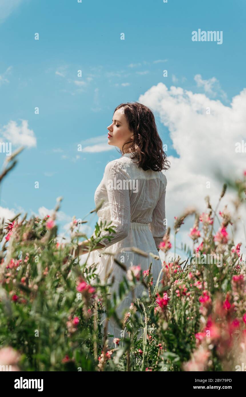 Seitenansicht Porträt einer jungen Frau in einem langen weißen Kleid stehen mit einem wilden Blumenstrauß in einem blühenden Blumenfeld gegen schöne helle blau Stockfoto