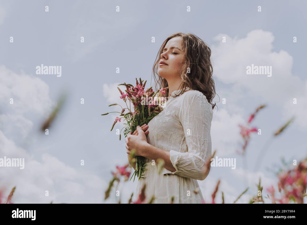 Mittelkörper Porträt einer jungen Frau in weißem Kleid mit geschlossenen Augen, hält einen Blumenstrauß, gegen blauen hellen Himmel geschossen Stockfoto