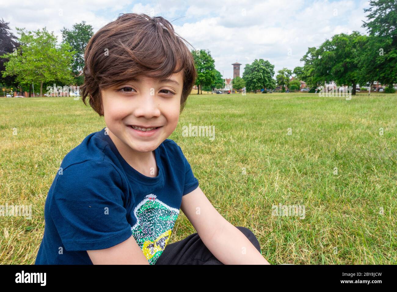 Ein kleiner Junge sitzt auf dem Boden im Park, lächelt und posiert für ein Porträt. Stockfoto