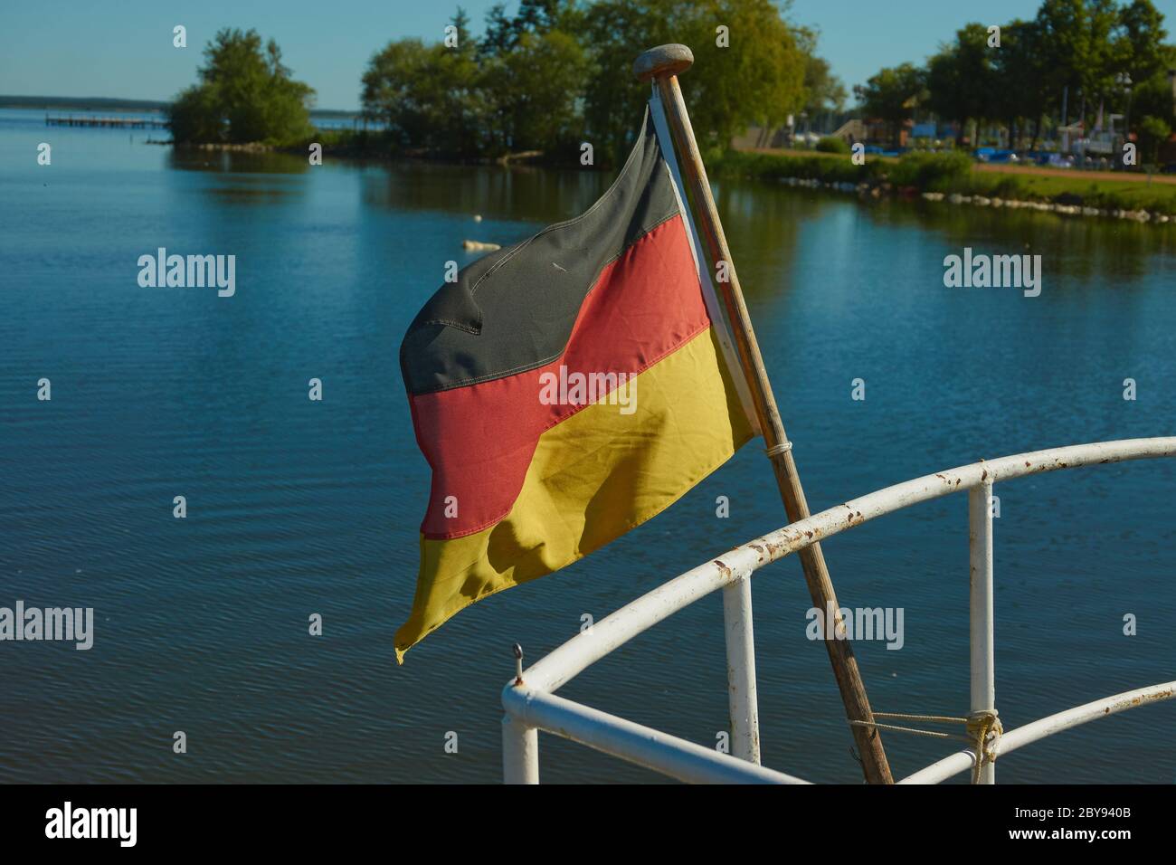 Schwarz-rot-goldene Flagge Deutschlands auf dem Heck eines Schiffes auf einem blauen See mit grünen Büschen am Ufer Stockfoto