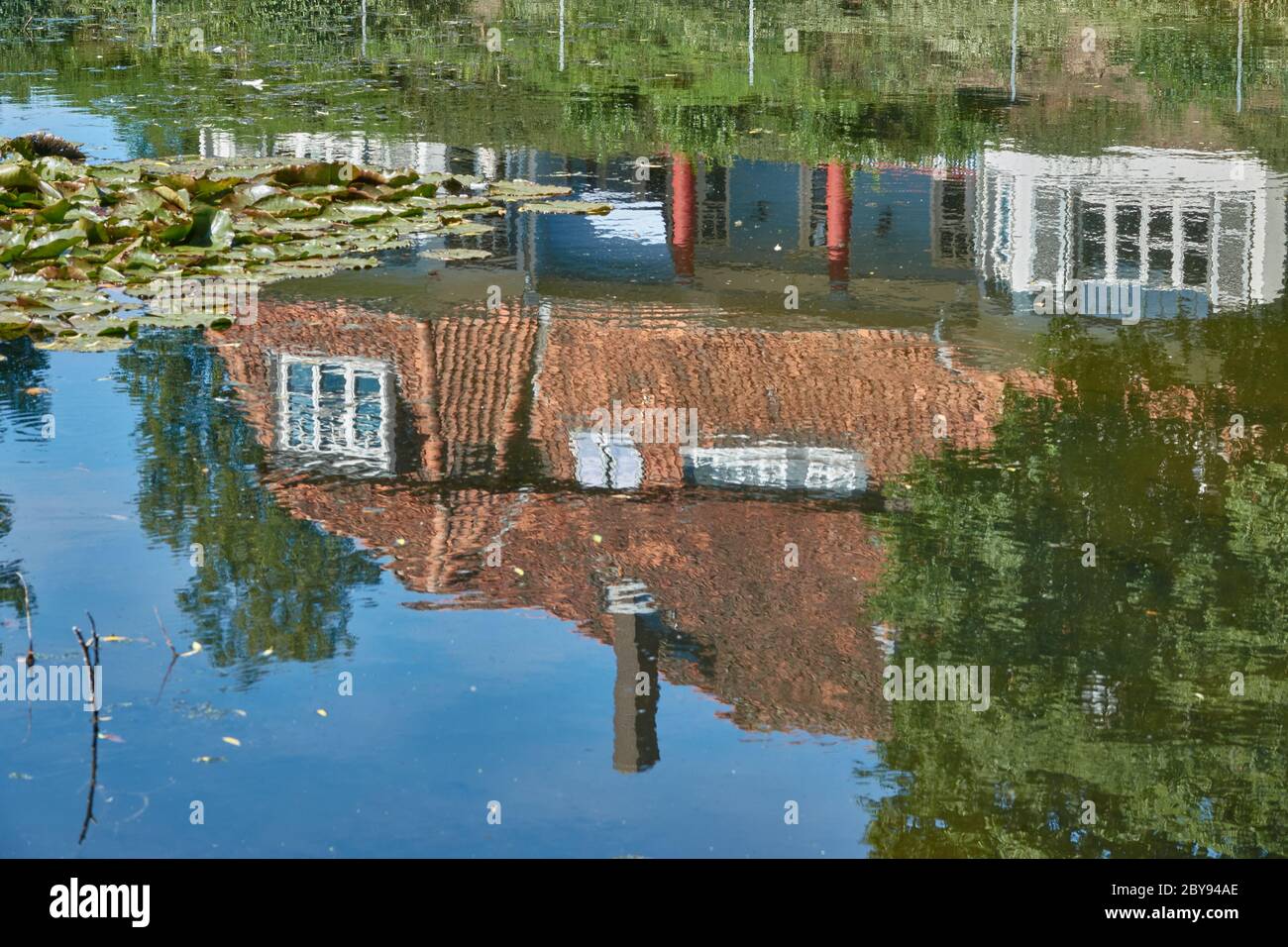 Spiegelung eines Hauses mit roten Dachziegeln und einem grünen Baum auf der Oberfläche eines Teiches mit Seerosenblättern Stockfoto