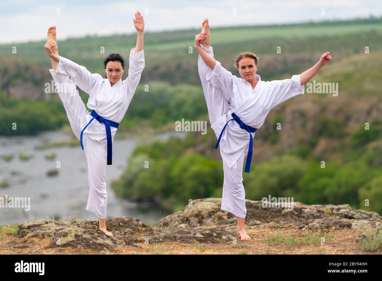 Zwei junge Frau, die Acro Yoga zusammen in einer synchronisierten Pose ausbalanciert auf einem Bein in einer vollen Strecke im Freien mit Blick auf ein Tal und Fluss Stockfoto