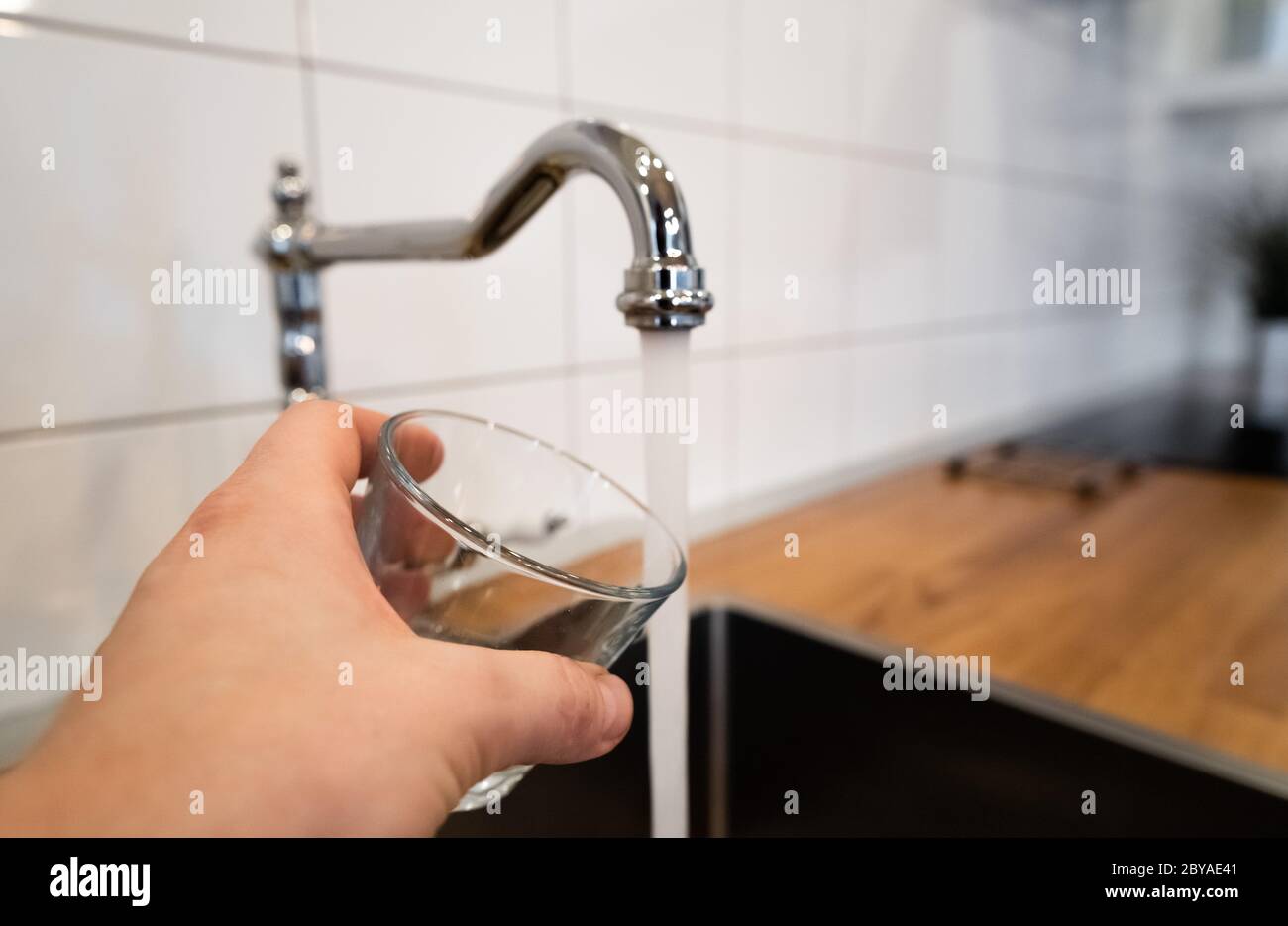 mans Hand hält ein leeres Glas in der Nähe einer Edelstahl-Küchenarmatur. Person erwägen Ausgießen von fließendem Wasser aus Chrom Wasserhahn. Sicher zu trinken oder nicht Stockfoto