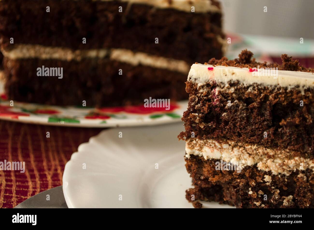 Ein Stück Schokoladenkuchen mit Sahne auf einem weißen Teller vor dem Hintergrund des restlichen Kuchens auf einer burgunderroten Tischdecke Stockfoto