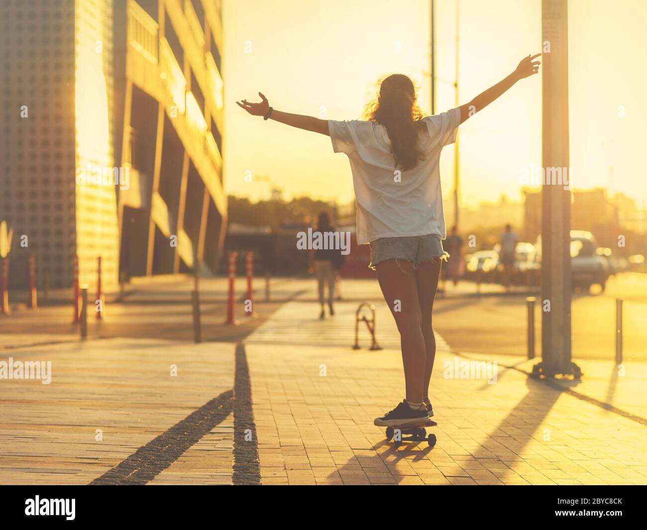 Mädchen auf einem Skateboard von den Strahlen der untergehenden Sonne beleuchtet, mit hoch erhobenen Armen, ihren Rücken zur Kamera. Selektiver Fokus. Stockfoto