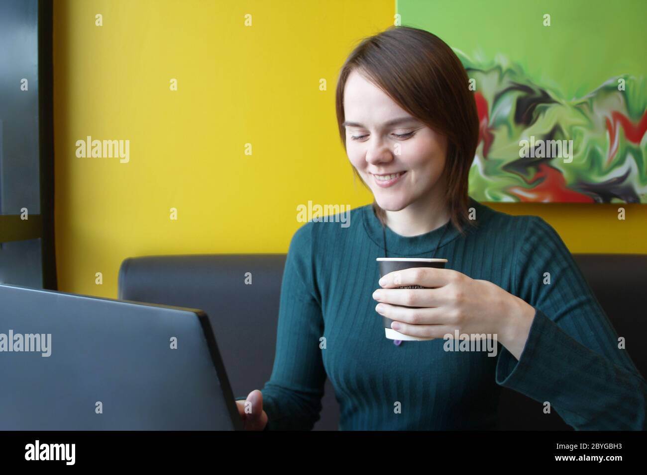 Ein Mädchen mit einem fröhlichen Gesichtsausdruck lächelt, während es an einem Laptop an einem Tisch in einem Café auf einem braunen Sofa gegen eine gelbe Wand sitzt. Er schaut auf einen Computermonitor und hält eine Papptasse mit Kaffee in der Hand. Stockfoto