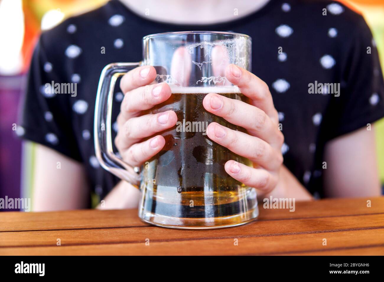 Nahaufnahme von weiblichen Händen, die einen großen, bedeckten Glaskrug mit kaltem Bier an einem Holztisch in einem Café halten. Erfrischendes Sommergetränk. Stockfoto