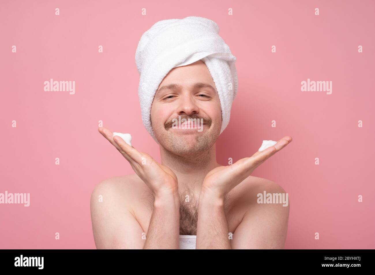 Schöner lustiger Mann mit Handtuch auf dem Kopf, der seine Haut mit Schaum tut Spa-Hautpflege. Studio auf rosa Wand gedreht. Stockfoto