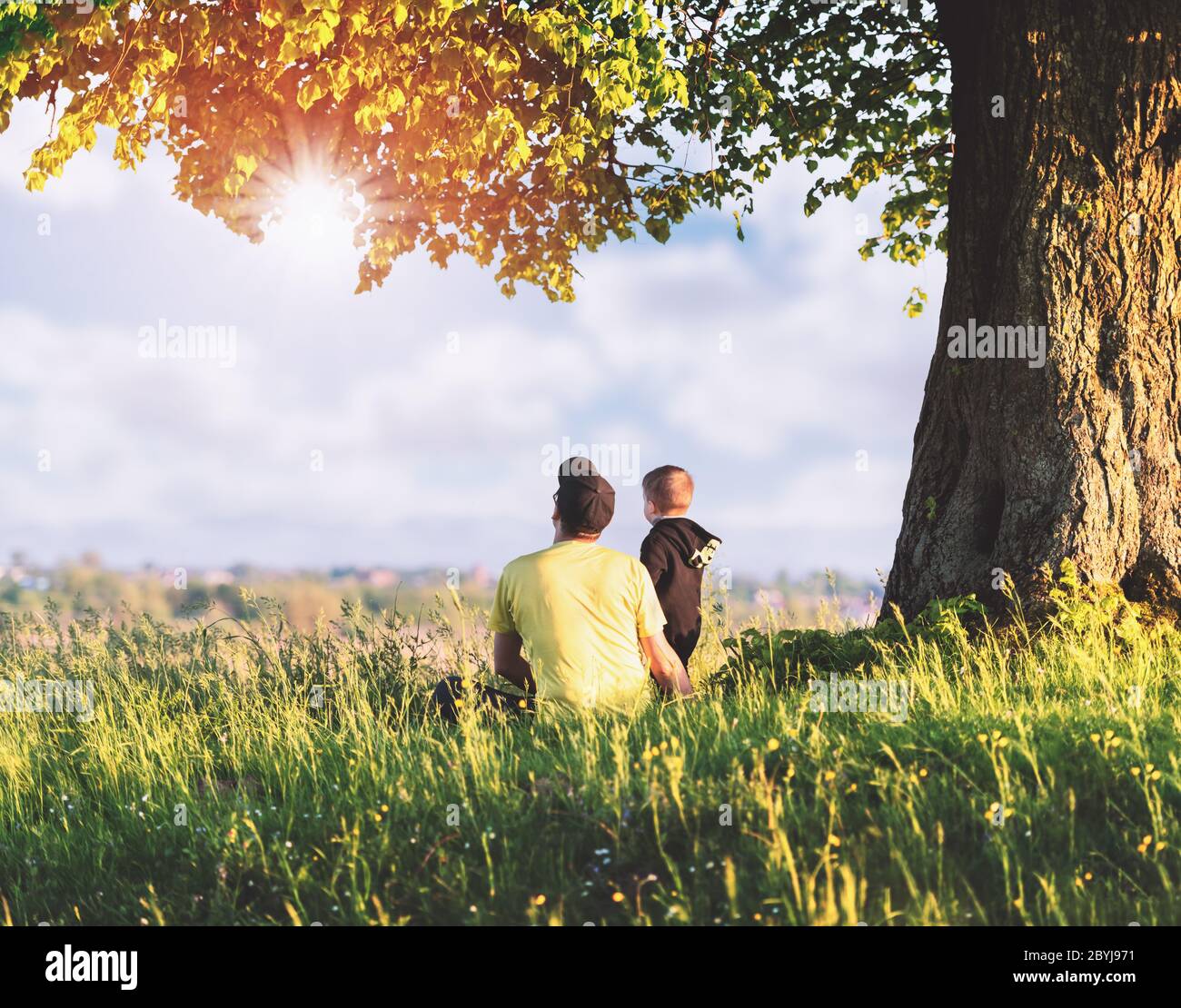 Vater mit Sohn auf der Frühlingswiese, der unter dem Baum in hohem Gras sitzt. Reisen mit Kind Konzept Stockfoto