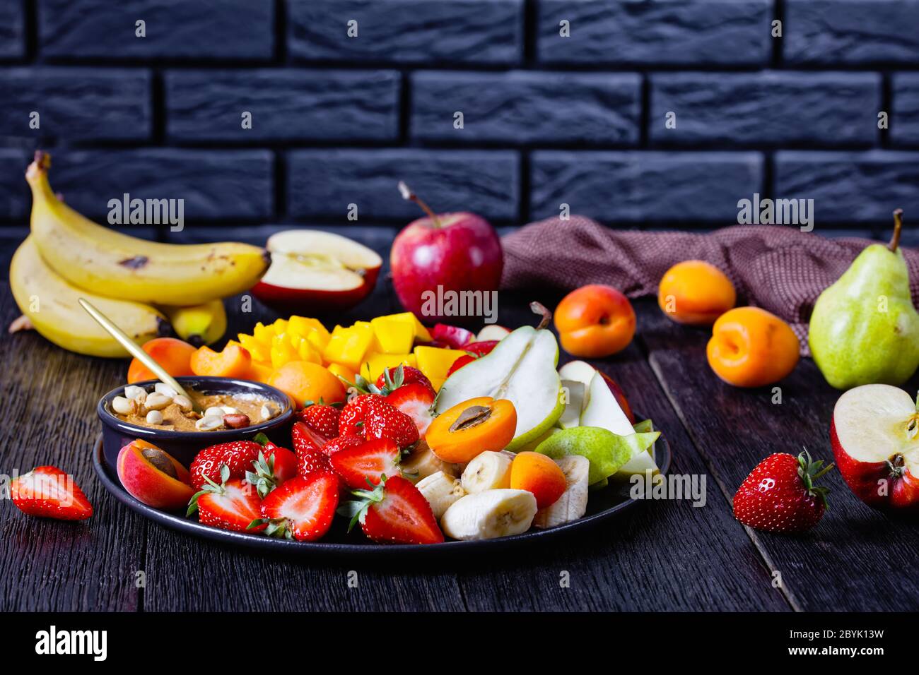 Fruchtmix mit Erdnussbutter Dip: Erdbeeren, tropische Mango, Banane, Äpfel, Birnen, Aprikosen und Erdnüsse auf einem schwarzen Teller auf einem dunklen Holzrückerich Stockfoto