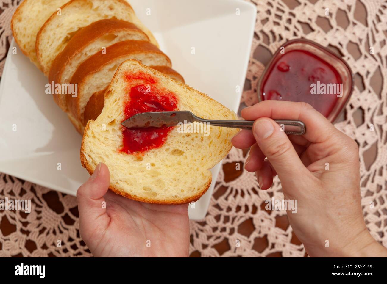 Traditionelles brasilianisches hausgemachtes Brot - Hände, die Erdbeermarmelade auf Brot legen. Häkelhandtuch Hintergrund. Nahaufnahme. Draufsicht. Stockfoto