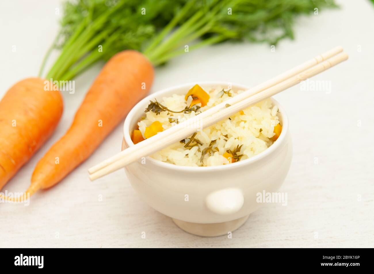 Vegetarische / vegane Küche - Rohe Karotten mit Blättern und eine Schüssel gekochten Reis mit Karotten. Essstäbchen oben auf dem Behälter. Stockfoto