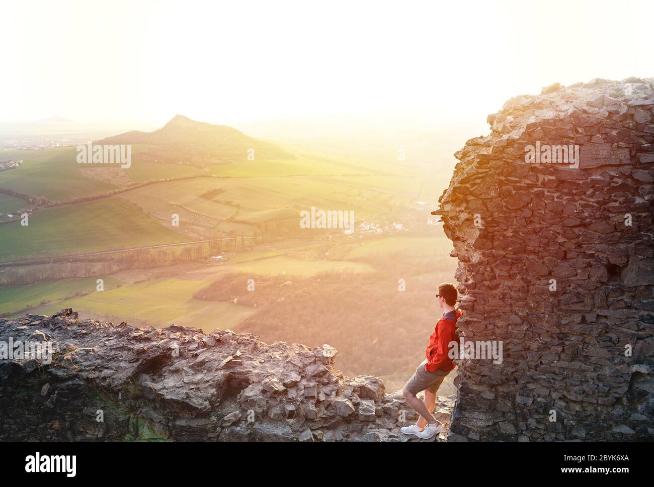 Professioneller Mann sitzt auf Burgruinen in aktiver Kleidung auf dem Gipfel des Berges ruhen und bewundern grüne Hügel und atemberaubende Landschaft Stockfoto