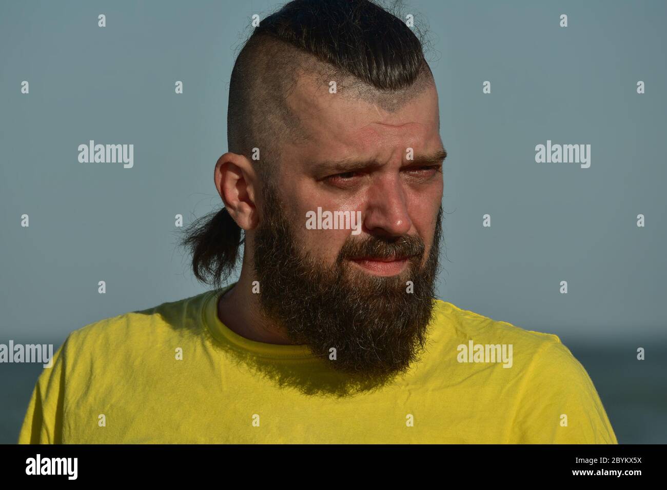 Ein junger Mann, der ein gelbes T-Shirt und Shorts trägt. Mann in einem gelben T-Shirt mit Bart auf dem Hintergrund des Meeres. Stockfoto