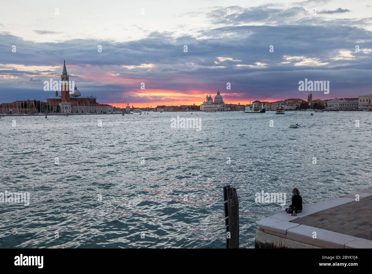 Die einsame Dame saß am Kai entlang Riva degli Schiavoni, Venedig, Italien und blickte in einen wunderschönen Sonnenuntergang über der Basilica di Santa Maria della Salute Stockfoto