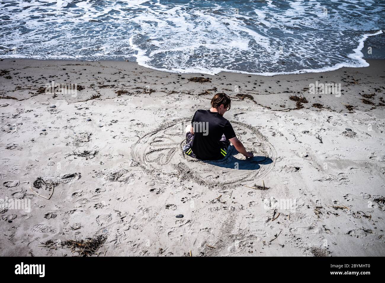 Dieser Mann sitzt am Strand und zieht im Sand seinen ,Kreis seines Lebens'. Die großen Herzen, die kleinen Kreise und die Bedeutung werden weggespült. Stockfoto