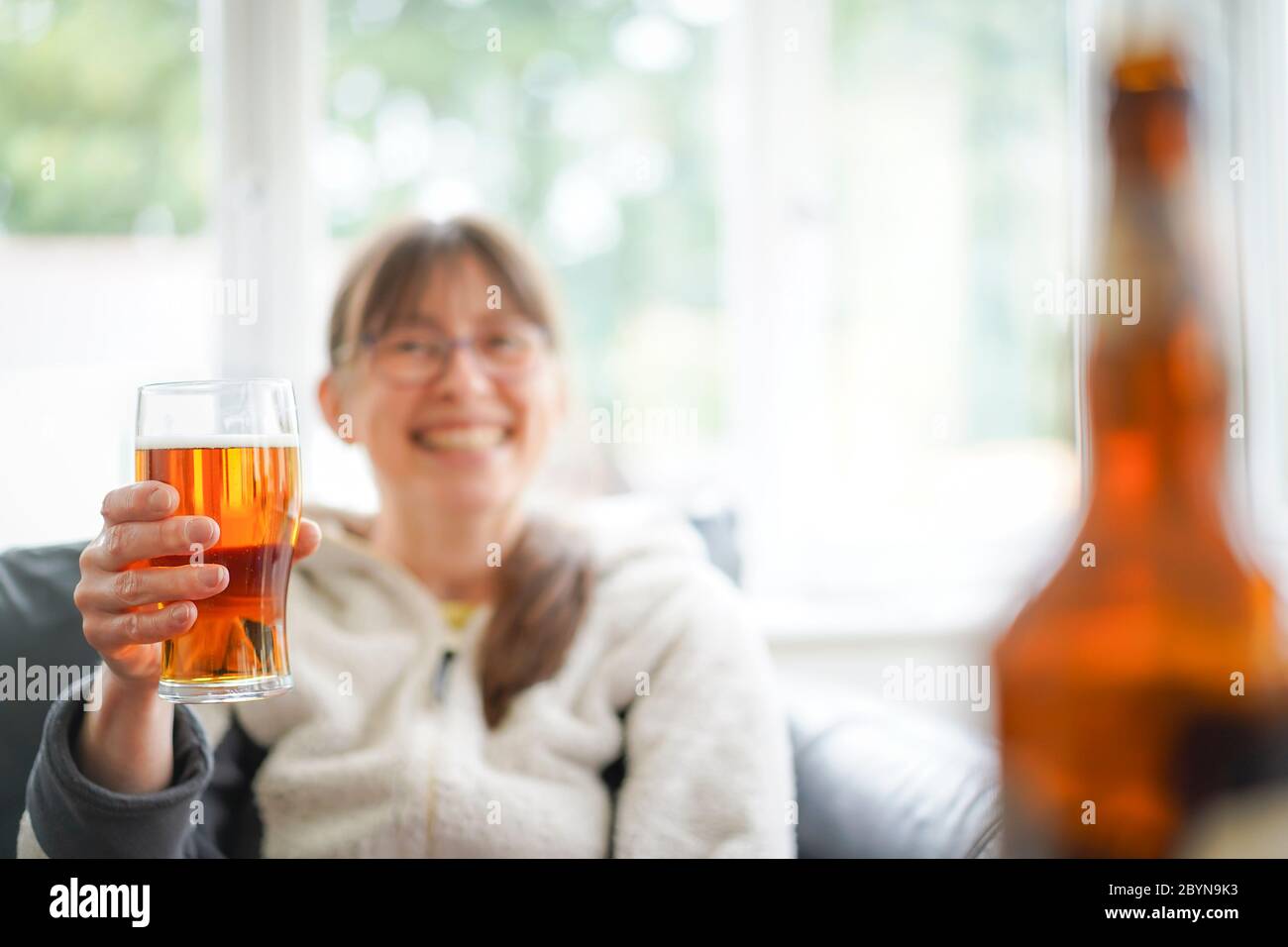 Isolierte junge Frau, die zu Hause Bier trinkt. Lady hält ein Pint Bier hoch und lächelt - Cheers! Stockfoto