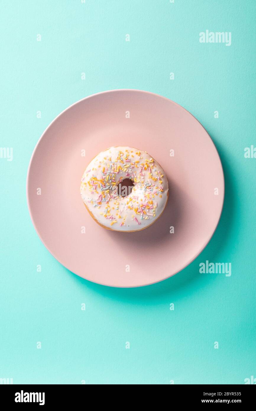 Vanilledonut mit Streuseln auf rosa Teller, süß glasierte Dessertspeise auf blauem minimalem Hintergrund, Draufsicht Stockfoto