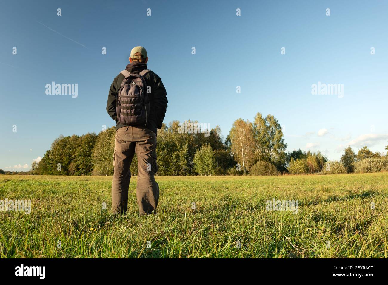 Ein Mann mit Rucksack auf einer grünen Wiese, schaut auf den Wald und den blauen Himmel, an einem sonnigen Tag gedreht Stockfoto