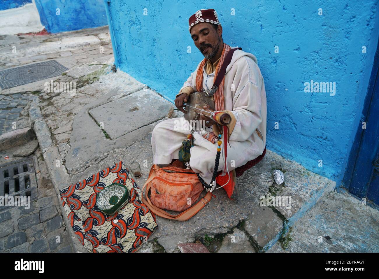 Der Straßenmusiker, der sich an der blauen Wand der Medina Rabats aufragt, spielt die dreisaitige Sintir, auch bekannt als Gimbri oder hejhouj, eine Hautfarbe Stockfoto