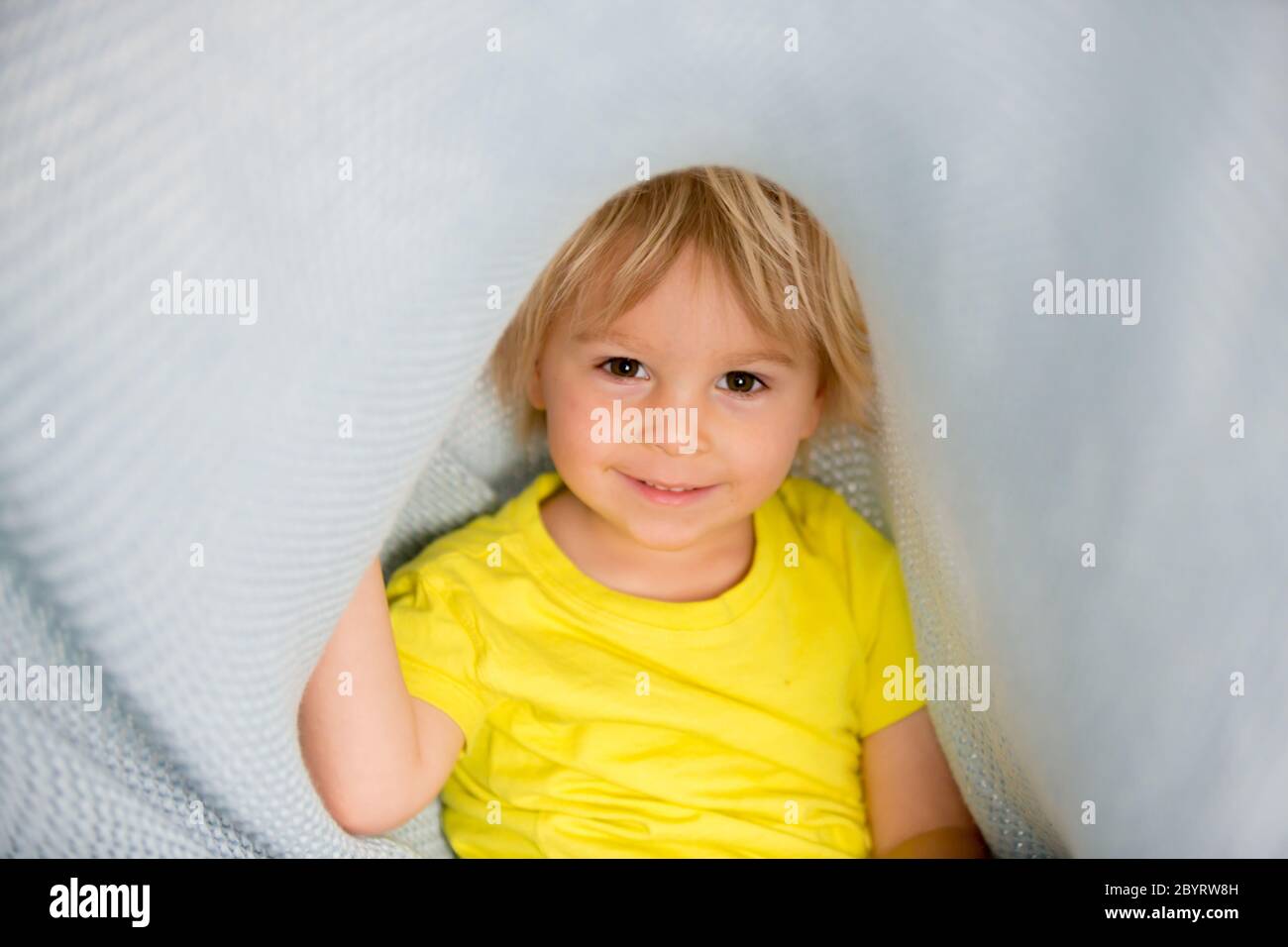 Kleiner kleiner kleiner kleiner Junge mit gelbem Hemd, versteckt unter Decke, lächelnd glücklich Stockfoto