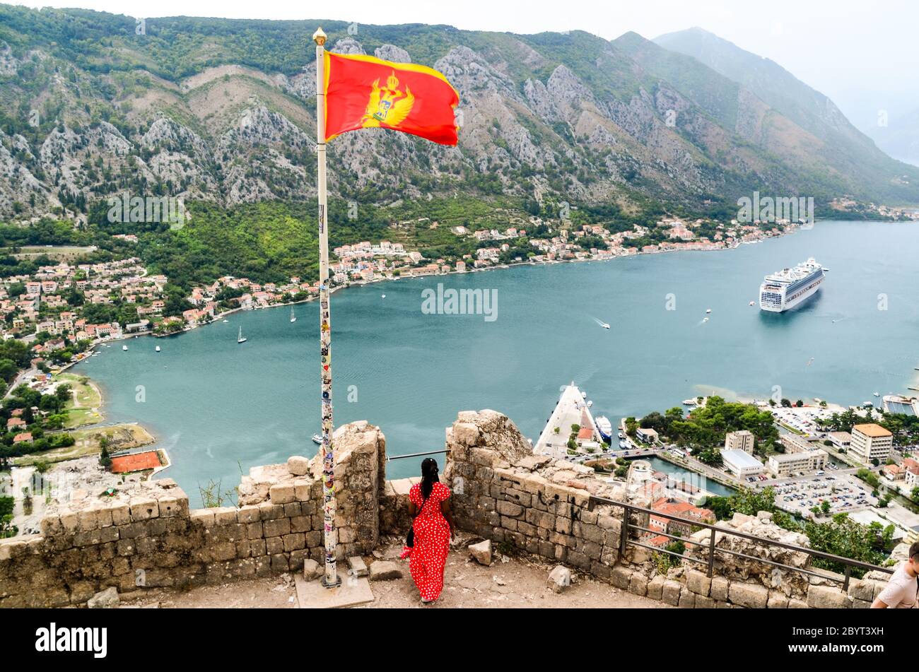 Frau in einem roten Kleid mit der roten Flagge Montenegros mit Blick auf ein großes Schiff in der Bucht von Kotor, Montenegro Stockfoto