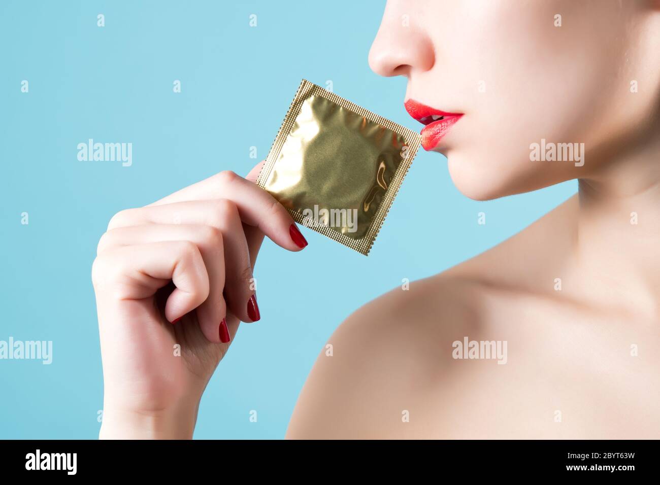 Der offene Mund des Mädchens mit rotem Lippenstift berührt ein Kondom In einer goldenen Verpackung Stockfoto