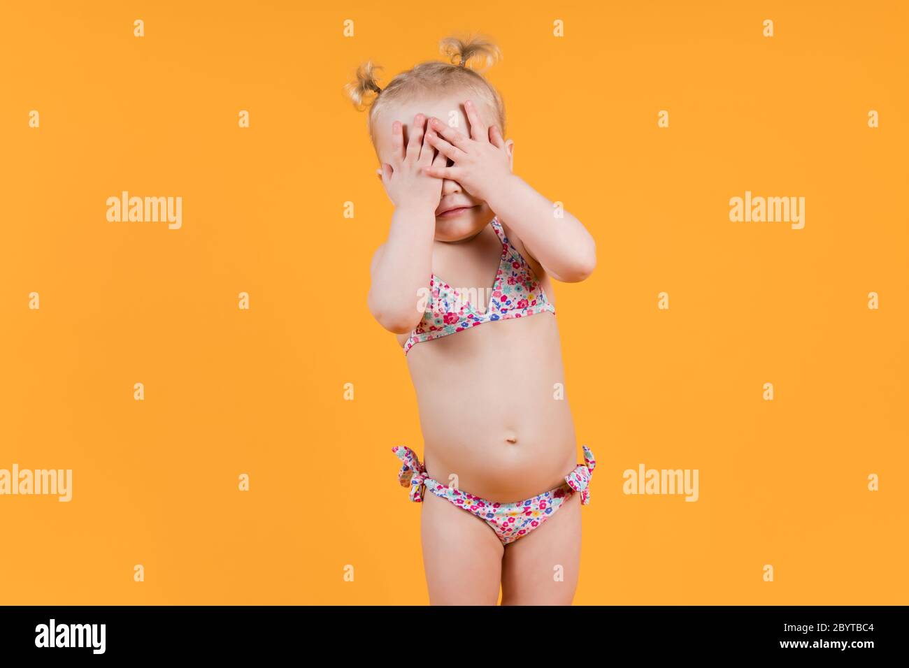 Ein kleines Mädchen in einem hellen Badeanzug bedeckt ihr Gesicht mit ihren Händen. Isoliert Stockfoto