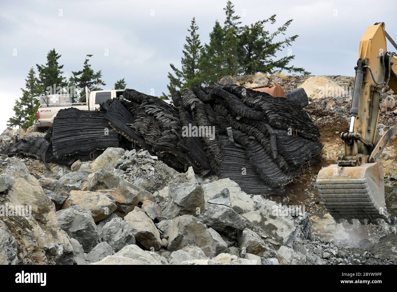 Schwarze recycelte Gummistrahlmatten (oben links) sitzen zwischen Felsen und gestrahltem Fels, während ein CAT (Caterpillar) Markenbagger einige der gestrahlten Ro reinigt Stockfoto
