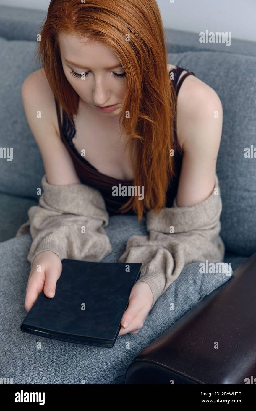 Rothaariges Mädchen, das auf einem Sofa sitzt und den Kopf über ein schwarzes Buch in den Händen beugt, Rahmen von oben. Stockfoto