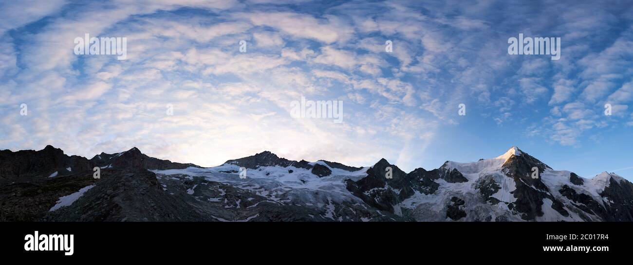 Panorama-Schnappschuss eines Bergrückens in der Schweiz am Morgen ist blauer Himmel mit welligen Wolken über verschneiten Gipfeln, aufgehende Sonne scheint am Ober Gabelhorn Stockfoto