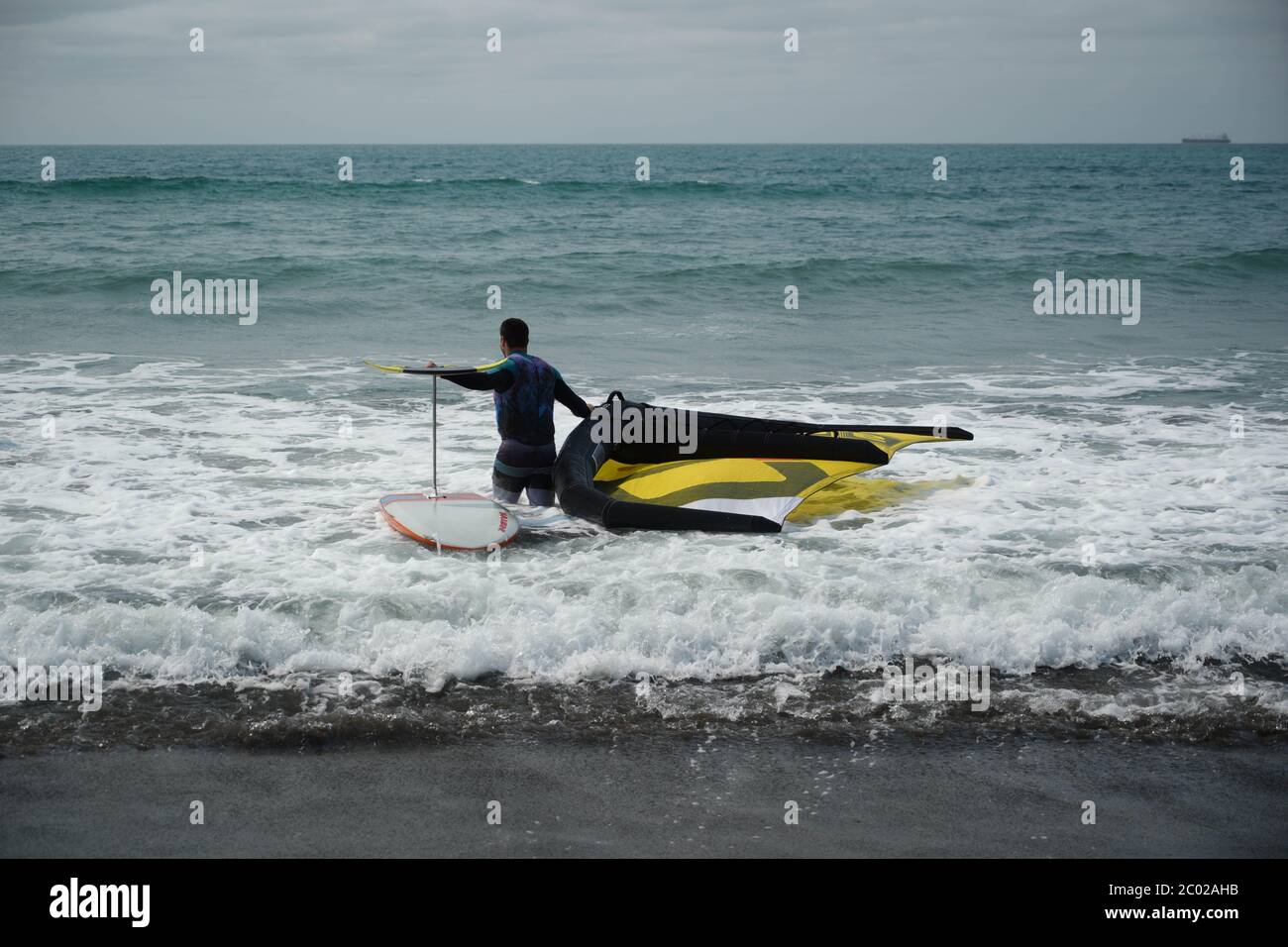 Ein Mann mit einem Neoprenanzug betritt das Meer mit einem handgehaltenen Flügel und einem Tragflügelbrett aus Wingfoil. Stockfoto